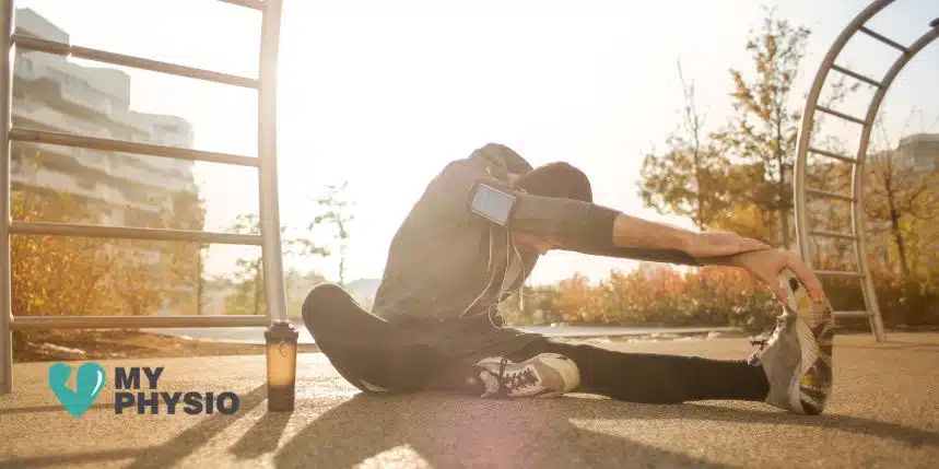 Man stretching outdoors in morning sunlight at an urban park, dressed in athletic wear with a smartphone armband and water bottle nearby, positioned near playground equipment, promoting physical therapy, injury prevention, and wellness with My Physio logo in the bottom left corner.